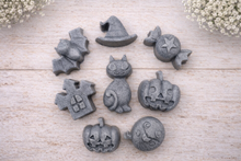 Load image into Gallery viewer, A collection of eight handmade vegan Halloween soaps featuring a bat, witch's hat, star-patterned sweet, haunted house, black cat, jack-o'-lantern, and a ghost, arranged on a light wooden surface with white floral accents.
