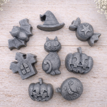 Load image into Gallery viewer, A collection of eight handmade vegan Halloween soaps featuring a bat, witch's hat, star-patterned sweet, haunted house, black cat, jack-o'-lantern, and a ghost, arranged on a light wooden surface with white floral accents.