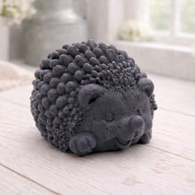 Load image into Gallery viewer, A detailed handmade vegan soap sculpted in the shape of a round, sitting hedgehog with intricate quill textures and a cute face, in a natural honey-brown tone, displayed on a light wooden surface.