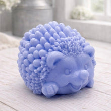 Load image into Gallery viewer, A detailed handmade vegan soap sculpted in the shape of a round, sitting hedgehog with intricate quill textures and a cute face, in a natural honey-brown tone, displayed on a light wooden surface.