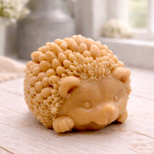Load image into Gallery viewer, A detailed handmade vegan soap sculpted in the shape of a round, sitting hedgehog with intricate quill textures and a cute face, in a natural honey-brown tone, displayed on a light wooden surface.