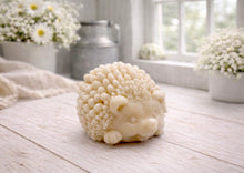 Load image into Gallery viewer, A detailed handmade vegan soap sculpted in the shape of a round, sitting hedgehog with intricate quill textures and a cute face, in a natural honey-brown tone, displayed on a light wooden surface.