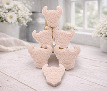 Load image into Gallery viewer, A collection of six handmade vegan soaps each intricately sculpted into the head of a shaggy Highland cow with long horns and a thick, textured coat, arranged in a pyramid on and around a small wooden easel.