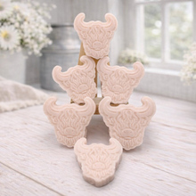 Load image into Gallery viewer, A collection of six handmade vegan soaps each intricately sculpted into the head of a shaggy Highland cow with long horns and a thick, textured coat, arranged in a pyramid on and around a small wooden easel.