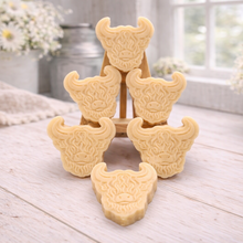 Load image into Gallery viewer, A collection of six handmade vegan soaps each intricately sculpted into the head of a shaggy Highland cow with long horns and a thick, textured coat, arranged in a pyramid on and around a small wooden easel.