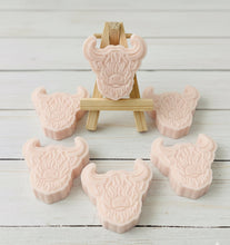 Load image into Gallery viewer, A collection of six handmade vegan soaps each intricately sculpted into the head of a shaggy Highland cow with long horns and a thick, textured coat, arranged in a pyramid on and around a small wooden easel.