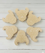 Load image into Gallery viewer, A collection of six handmade vegan soaps each intricately sculpted into the head of a shaggy Highland cow with long horns and a thick, textured coat, arranged in a pyramid on and around a small wooden easel.