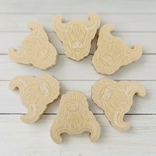 Load image into Gallery viewer, A collection of six handmade vegan soaps each intricately sculpted into the head of a shaggy Highland cow with long horns and a thick, textured coat, arranged in a pyramid on and around a small wooden easel.