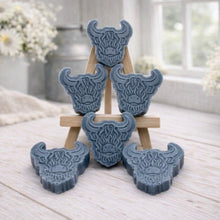 Load image into Gallery viewer, A collection of six handmade vegan soaps each intricately sculpted into the head of a shaggy Highland cow with long horns and a thick, textured coat, arranged in a pyramid on and around a small wooden easel.