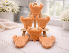 Load image into Gallery viewer, A collection of six handmade vegan soaps each intricately sculpted into the head of a shaggy Highland cow with long horns and a thick, textured coat, arranged in a pyramid on and around a small wooden easel.