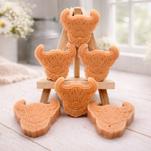 Load image into Gallery viewer, A collection of six handmade vegan soaps each intricately sculpted into the head of a shaggy Highland cow with long horns and a thick, textured coat, arranged in a pyramid on and around a small wooden easel.