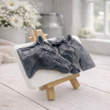 Load image into Gallery viewer, A rectangular handmade vegan soap featuring a detailed 3D relief of two horse heads leaning affectionately towards each other, finished in a slate-grey tone against a white soap base and displayed on a miniature wooden easel.