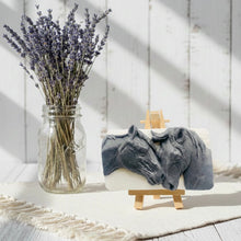 Load image into Gallery viewer, A grey and white handmade vegan soap featuring a highly detailed relief of two horses nuzzling heads, displayed on a wooden easel against a whimsical farmhouse decor background.