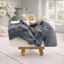 Load image into Gallery viewer, A rectangular handmade vegan soap featuring a detailed relief of two horse heads leaning affectionately towards each other, finished in a slate-grey tone against a white soap base and displayed on a miniature wooden easel.