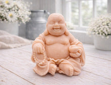 Load image into Gallery viewer, A 3D handmade vegan soap sculpted into a joyful Laughing Buddha sitting in a meditative pose, holding prayer beads and a small bowl, displayed in a bright farmhouse setting