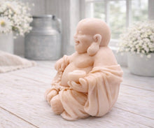 Load image into Gallery viewer, A 3D handmade vegan soap sculpted into a joyful Laughing Buddha sitting in a meditative pose, holding prayer beads and a small bowl, displayed in a bright farmhouse setting