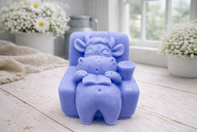 Load image into Gallery viewer, A handmade vegan soap sculpted to show a whimsical "lazy cow" character relaxing in an armchair, reading a book and holding a drink, displayed on a light wooden surface.