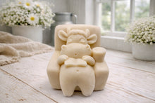 Load image into Gallery viewer, A handmade vegan soap sculpted to show a whimsical "lazy cow" character relaxing in an armchair, reading a book and holding a drink, displayed on a light wooden surface.
