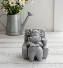 Load image into Gallery viewer, A handmade vegan soap sculpted to show a whimsical "lazy cow" character relaxing in an armchair, reading a book and holding a drink, displayed on a light wooden surface.
