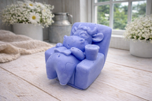 Load image into Gallery viewer, A handmade vegan soap sculpted to show a whimsical "lazy cow" character relaxing in an armchair, reading a book and holding a drink, displayed on a light wooden surface.