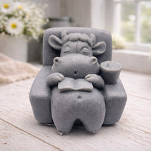 Load image into Gallery viewer, A handmade vegan soap sculpted to show a whimsical "lazy cow" character relaxing in an armchair, reading a book and holding a drink, displayed on a light wooden surface.