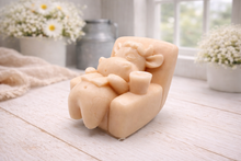 Load image into Gallery viewer, A handmade vegan soap sculpted to show a whimsical "lazy cow" character relaxing in an armchair, reading a book and holding a drink, displayed on a light wooden surface.