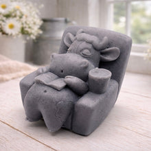 Load image into Gallery viewer, A handmade vegan soap sculpted to show a whimsical "lazy cow" character relaxing in an armchair, reading a book and holding a drink, displayed on a light wooden surface.
