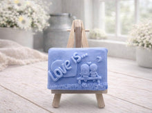 Load image into Gallery viewer, A rectangular handmade vegan soap featuring a detailed 3D relief carving of a young couple sitting on a bench under a crescent moon, with the words "Love is..." embossed inside a heart, displayed on a wooden easel.