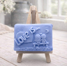 Load image into Gallery viewer, A rectangular handmade vegan soap featuring a detailed 3D relief carving of a young couple sitting on a bench under a crescent moon, with the words "Love is..." embossed inside a heart, displayed on a wooden easel.
