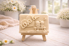 Load image into Gallery viewer, A rectangular handmade vegan soap featuring a detailed 3D relief carving of a young couple sitting on a bench under a crescent moon, with the words "Love is..." embossed inside a heart, displayed on a wooden easel.
