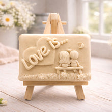 Load image into Gallery viewer, A rectangular handmade vegan soap featuring a detailed 3D relief carving of a young couple sitting on a bench under a crescent moon, with the words "Love is..." embossed inside a heart, displayed on a wooden easel.