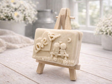 Load image into Gallery viewer, A rectangular handmade vegan soap featuring a detailed 3D relief carving of a young couple sitting on a bench under a crescent moon, with the words "Love is..." embossed inside a heart, displayed on a wooden easel.