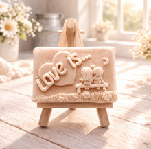 Load image into Gallery viewer, A rectangular handmade vegan soap featuring a detailed 3D relief carving of a young couple sitting on a bench under a crescent moon, with the words "Love is..." embossed inside a heart, displayed on a wooden easel.