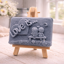 Load image into Gallery viewer, A rectangular handmade vegan soap featuring a detailed 3D relief carving of a young couple sitting on a bench under a crescent moon, with the words "Love is..." embossed inside a heart, displayed on a wooden easel.