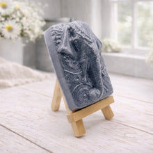 Load image into Gallery viewer, A rectangular handmade vegan soap in a speckled slate-grey finish, featuring a highly detailed relief of an elephant's head adorned with ornate, traditional patterns and a decorative floral background, displayed on a miniature wooden easel.