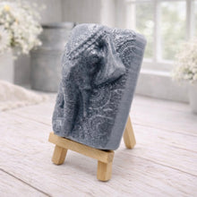 Load image into Gallery viewer, A rectangular handmade vegan soap in a speckled slate-grey finish, featuring a highly detailed relief of an elephant's head adorned with ornate, traditional patterns and a decorative floral background, displayed on a miniature wooden easel.