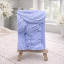 Load image into Gallery viewer, A rectangular handmade vegan soap featuring a high-relief carving of a mother tenderly cradling a small baby in her arms, displayed on a miniature wooden easel in a bright farmhouse setting.