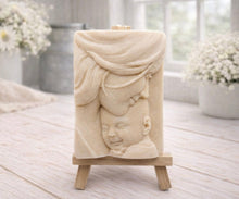 Load image into Gallery viewer, A rectangular handmade vegan soap featuring a high-relief carving of a mother tenderly cradling a small baby in her arms, displayed on a miniature wooden easel in a bright farmhouse setting.