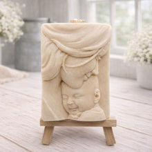 Load image into Gallery viewer, A rectangular handmade vegan soap featuring a high-relief carving of a mother tenderly cradling a small baby in her arms, displayed on a miniature wooden easel in a bright farmhouse setting.