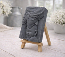 Load image into Gallery viewer, A rectangular handmade vegan soap featuring a high-relief carving of a mother tenderly cradling a small baby in her arms, displayed on a miniature wooden easel in a bright farmhouse setting.