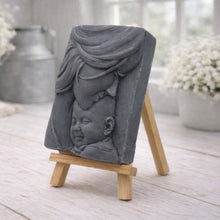 Load image into Gallery viewer, A rectangular handmade vegan soap featuring a high-relief carving of a mother tenderly cradling a small baby in her arms, displayed on a miniature wooden easel in a bright farmhouse setting.