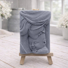 Load image into Gallery viewer, A rectangular handmade vegan soap featuring a high-relief carving of a mother tenderly cradling a small baby in her arms, displayed on a miniature wooden easel in a bright farmhouse setting.
