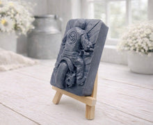 Load image into Gallery viewer, A rectangular handmade vegan soap featuring a detailed high-relief carving of a biker from behind riding a motorcycle, wearing a leather-style jacket with a skull emblem on the back, displayed on a miniature wooden easel.