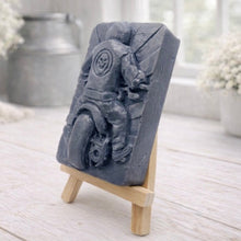 Load image into Gallery viewer, A rectangular handmade vegan soap featuring a detailed high-relief carving of a biker from behind riding a motorcycle, wearing a leather-style jacket with a skull emblem on the back, displayed on a miniature wooden easel.