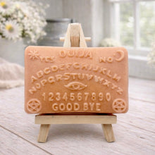 Load image into Gallery viewer, A rectangular handmade vegan soap featuring a highly detailed relief of a classic Ouija spirit board with the alphabet, numbers 0-9, an eye symbol, and the words "Yes," "No," and "Good Bye," displayed on a miniature wooden easel.