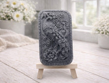 Load image into Gallery viewer, handmade vegan soap featuring a highly detailed relief carving of a peacock with a long, flowing tail, surrounded by delicate cherry blossoms and a large peony, displayed on a wooden easel.