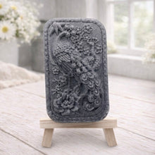 Load image into Gallery viewer, handmade vegan soap featuring a highly detailed relief carving of a peacock with a long, flowing tail, surrounded by delicate cherry blossoms and a large peony, displayed on a wooden easel.
