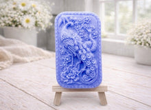 Load image into Gallery viewer, handmade vegan soap featuring a highly detailed relief carving of a peacock with a long, flowing tail, surrounded by delicate cherry blossoms and a large peony, displayed on a wooden easel.