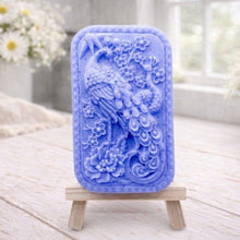 Load image into Gallery viewer, handmade vegan soap featuring a highly detailed relief carving of a peacock with a long, flowing tail, surrounded by delicate cherry blossoms and a large peony, displayed on a wooden easel.