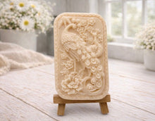 Load image into Gallery viewer, handmade vegan soap featuring a highly detailed relief carving of a peacock with a long, flowing tail, surrounded by delicate cherry blossoms and a large peony, displayed on a wooden easel.